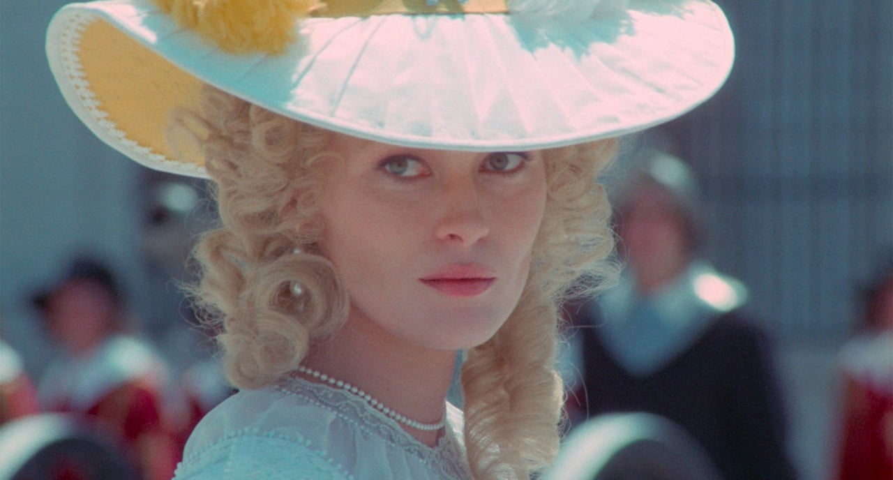 A woman in 18th-century attire with curled blonde hair and a large white hat adorned with yellow flowers gazes forward, with blurred figures in the background.