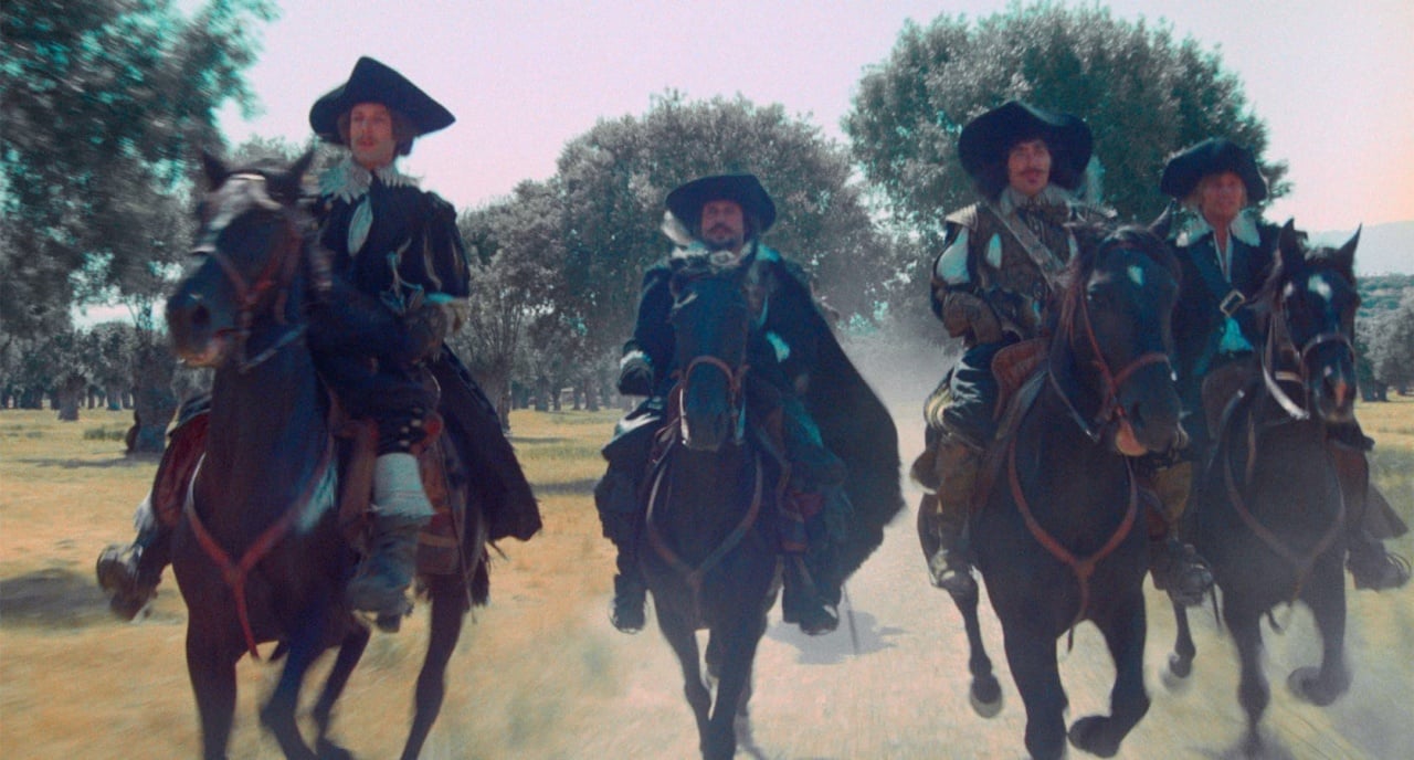 Four men in historical attire ride horses side by side across a sunlit field, with trees in the background.