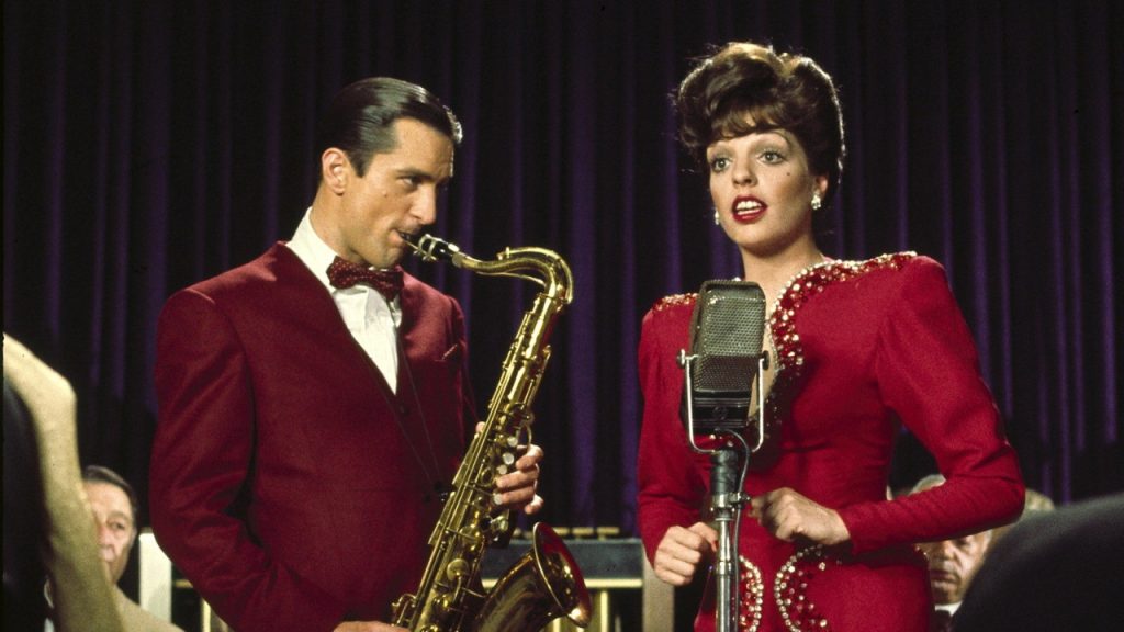 A man in a red suit plays a saxophone while a woman in a red dress sings into a vintage microphone on stage with a band in the background.