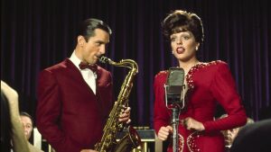 A man in a red suit plays a saxophone while a woman in a red dress sings into a vintage microphone on stage with a band in the background.