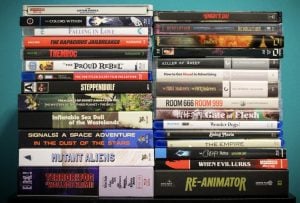 Stacked arrangement of DVDs and Blu-rays against a teal wall, featuring various movie titles across different genres.