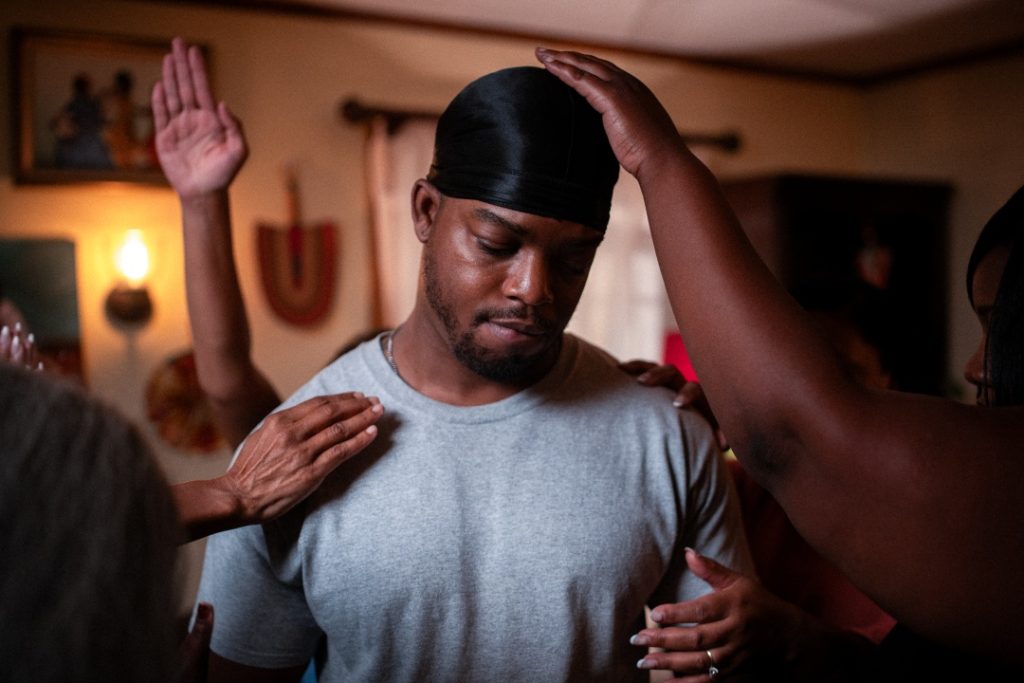 A man in a gray shirt stands with his eyes closed as several people place their hands on his head and shoulders in a dimly lit room.