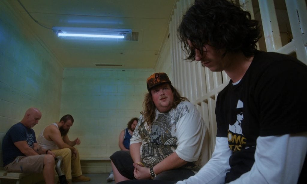 Five men sit on benches inside a jail cell, two of them engaged in conversation while the others look down or away.
