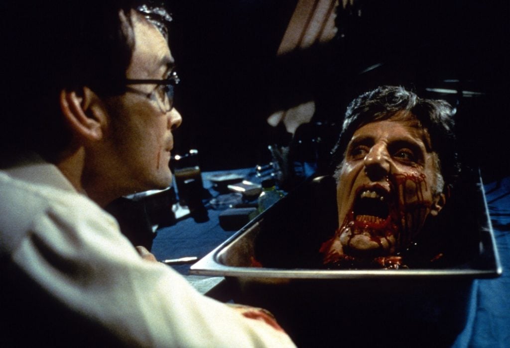 A man in glasses looks at a severed, reanimated head in a bloody tray on a table, with medical instruments and bottles in the background.