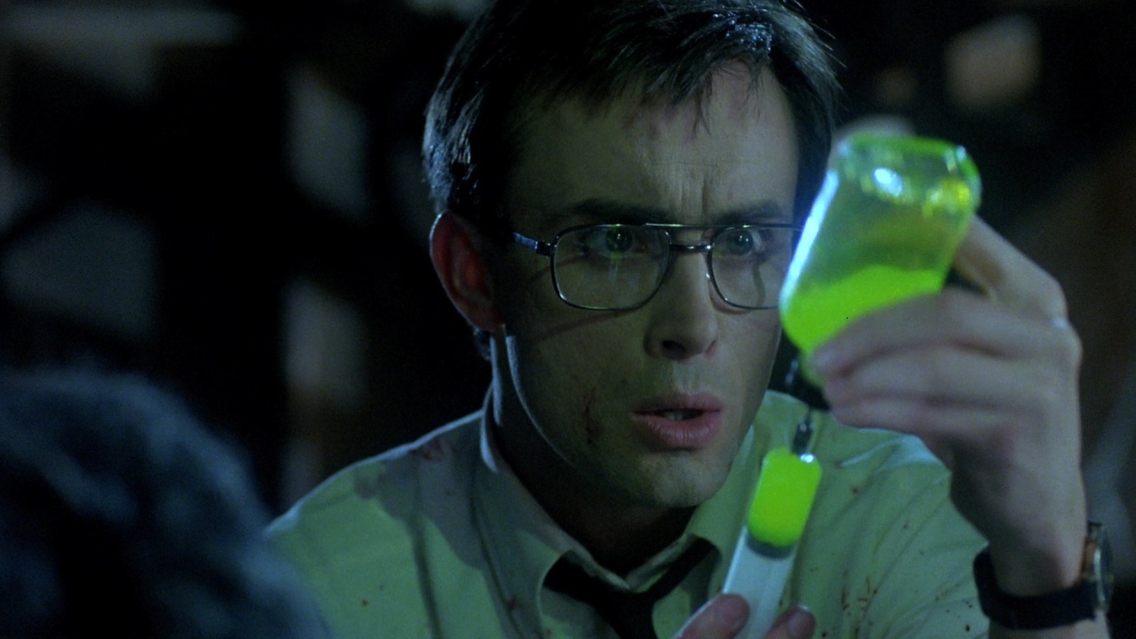 A man wearing glasses and a white shirt carefully fills a syringe with a glowing green liquid from a vial.