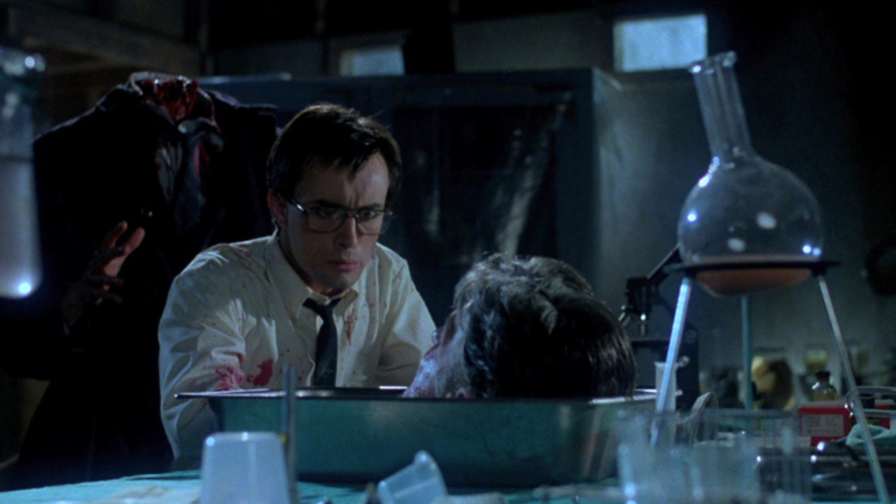 A man in glasses leans over a severed head in a metal tray on a laboratory table, with scientific glassware and a headless figure nearby.