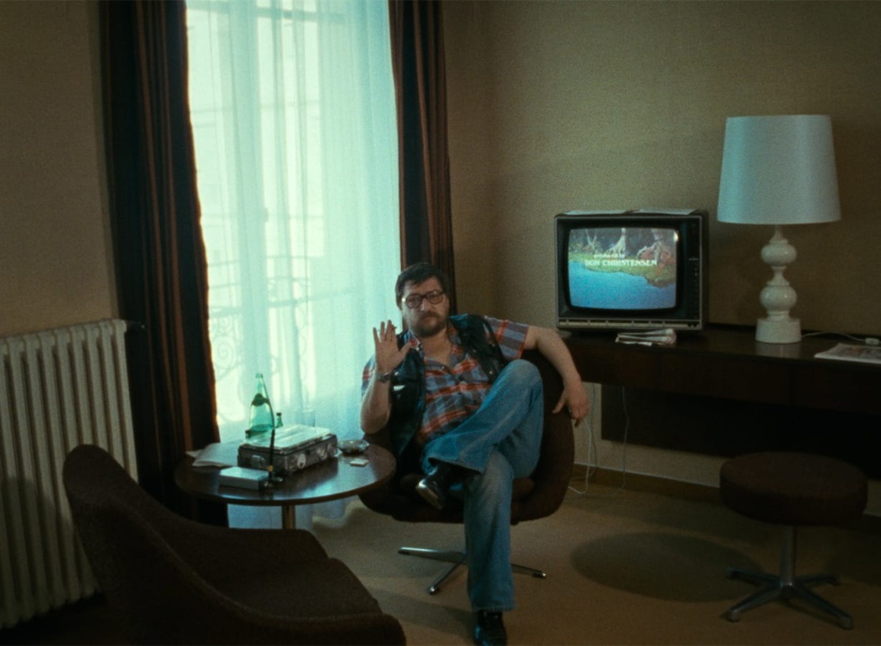 A man sits in a mid-century modern room, facing the camera, with a TV displaying "MERRY CHRISTMAS" in the background and a table holding books and a bottle.