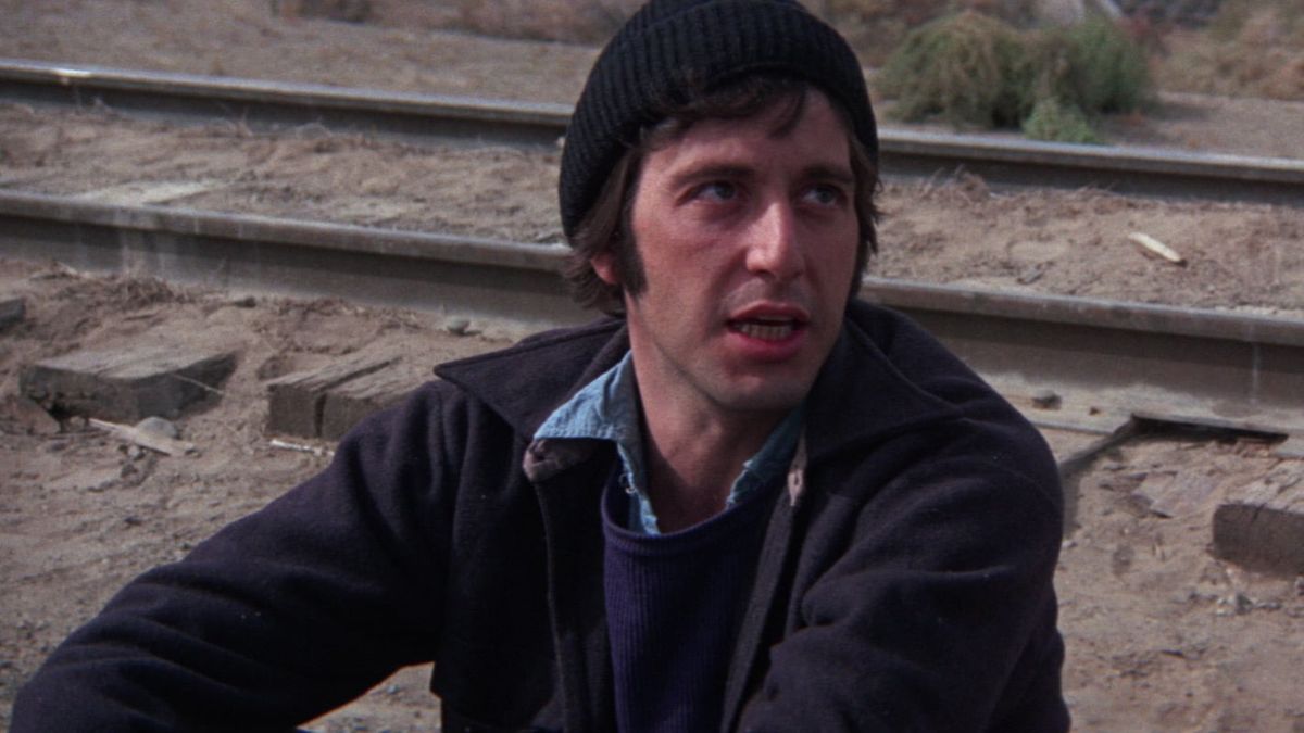 A man wearing a black beanie and dark jacket sits on the ground near railroad tracks, looking to the side.
