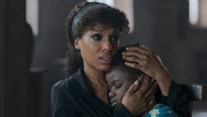 A woman sits holding a sleeping child in her arms, looking concerned while comforting the child in a dimly lit room.