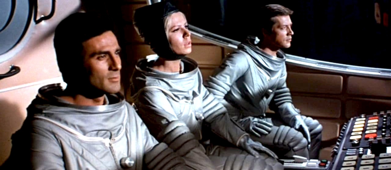 Three astronauts in silver spacesuits sit at a control panel, focused on their tasks inside a spacecraft cockpit.