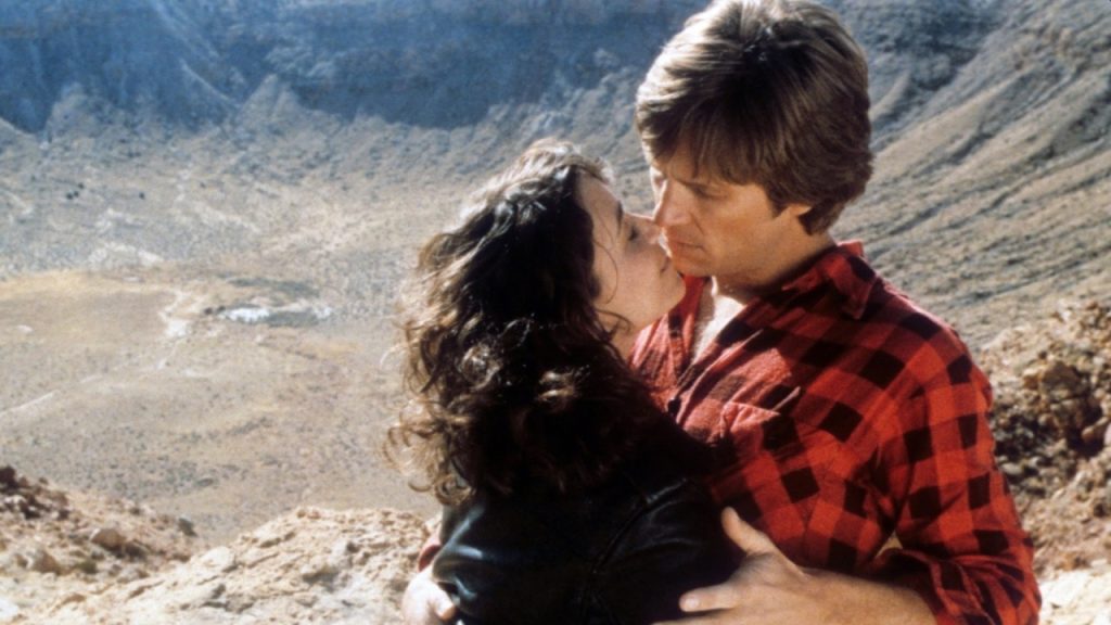 A man in a red plaid shirt and a woman in a black jacket embrace closely on rocky terrain with a canyon landscape in the background.