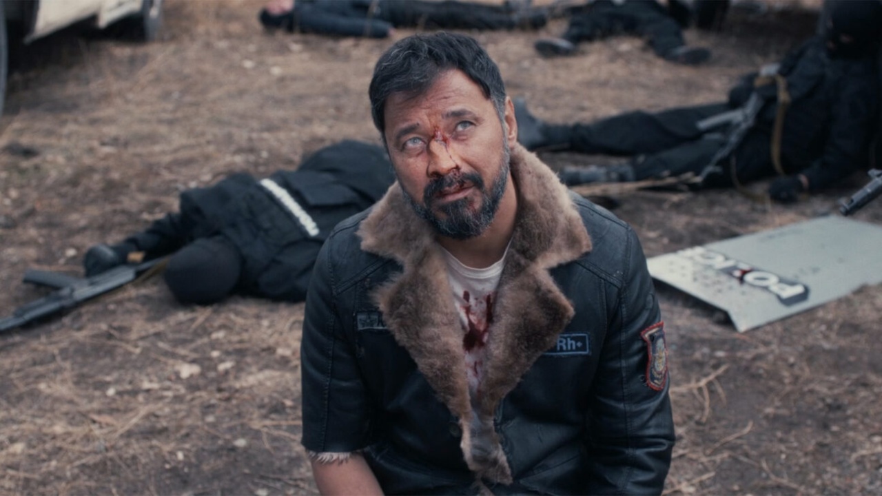 A man with blood on his chest and cloudy eyes kneels on the ground, surrounded by fallen people in dark uniforms on a dirt surface.
