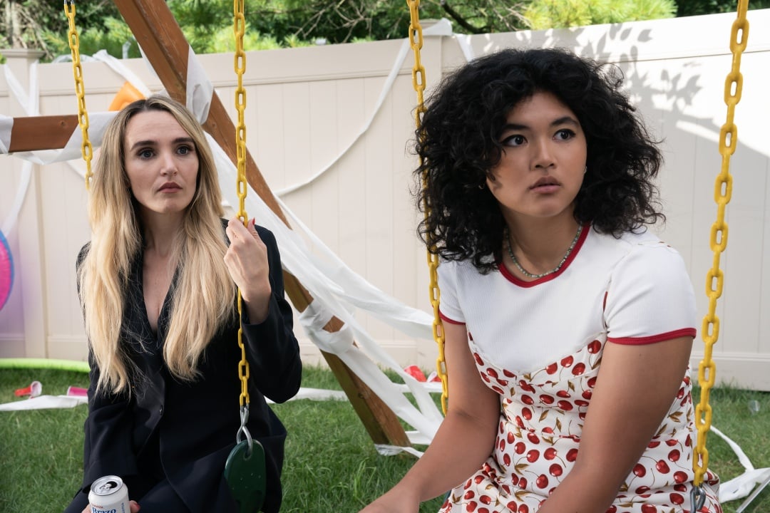 Two women sit on yellow swings in a backyard, with one holding a can and the other looking off to the side. A swing set and scattered items are visible in the background.