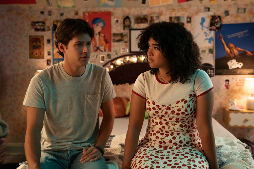 A young man and woman sit on a bed in a decorated bedroom, facing each other with serious expressions.