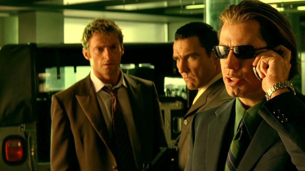 Three men in suits stand indoors; one in sunglasses talks on a phone while the other two look on with serious expressions.