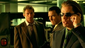 Three men in suits stand indoors; one in sunglasses talks on a phone while the other two look on with serious expressions.