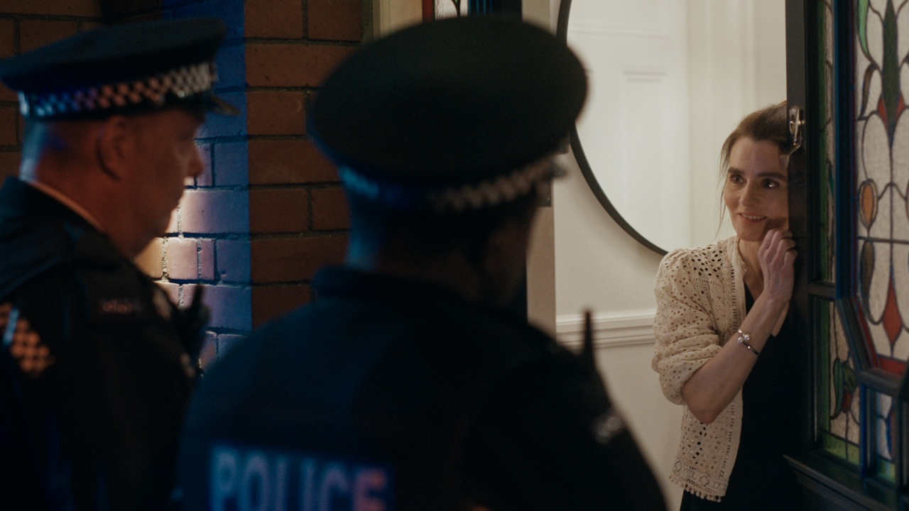 Two police officers stand at the door of a home, speaking with a woman who is partially hiding behind the door and looking at them.