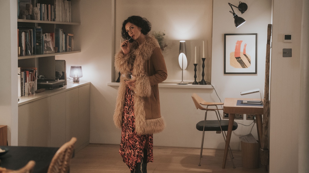 A woman in a fur-trimmed coat stands in a home office, talking on the phone; a desk, bookshelves, and framed art are visible in the background.