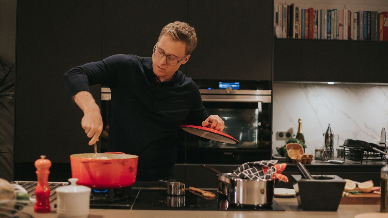 A person stirs food in a red pot on a modern kitchen stove, holding the pot lid in one hand. Various kitchen utensils and ingredients are on the countertop.