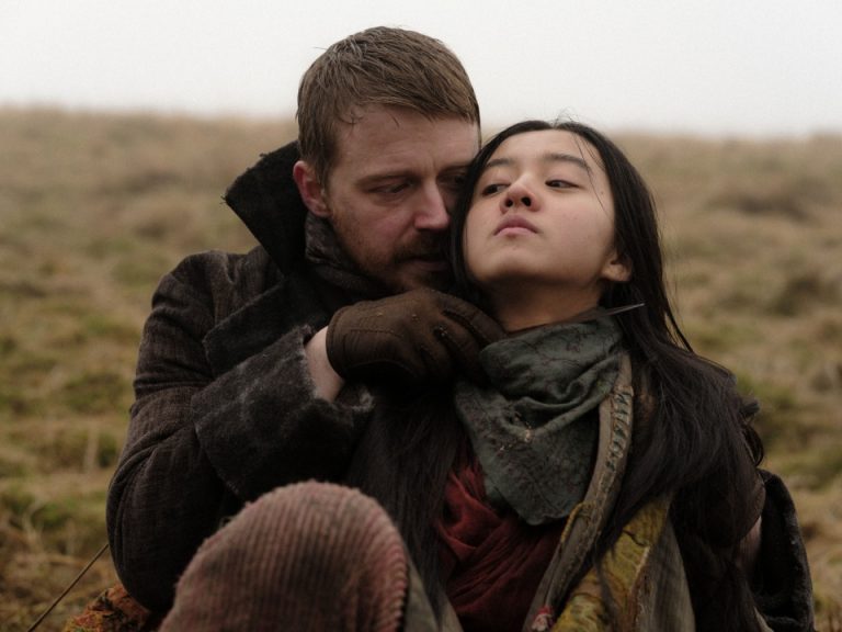 ‘Tornado’ 4K UHD Blu-Ray Review – A Slow Burn Tale Of Revenge A man in a brown coat and gloves holds a scarf around a woman’s neck as they sit together on a grassy field under a gray, overcast sky.