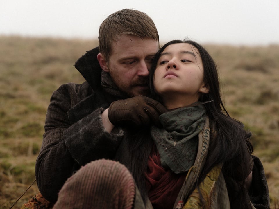 A man in a brown coat and gloves holds a scarf around a woman’s neck as they sit together on a grassy field under a gray, overcast sky.