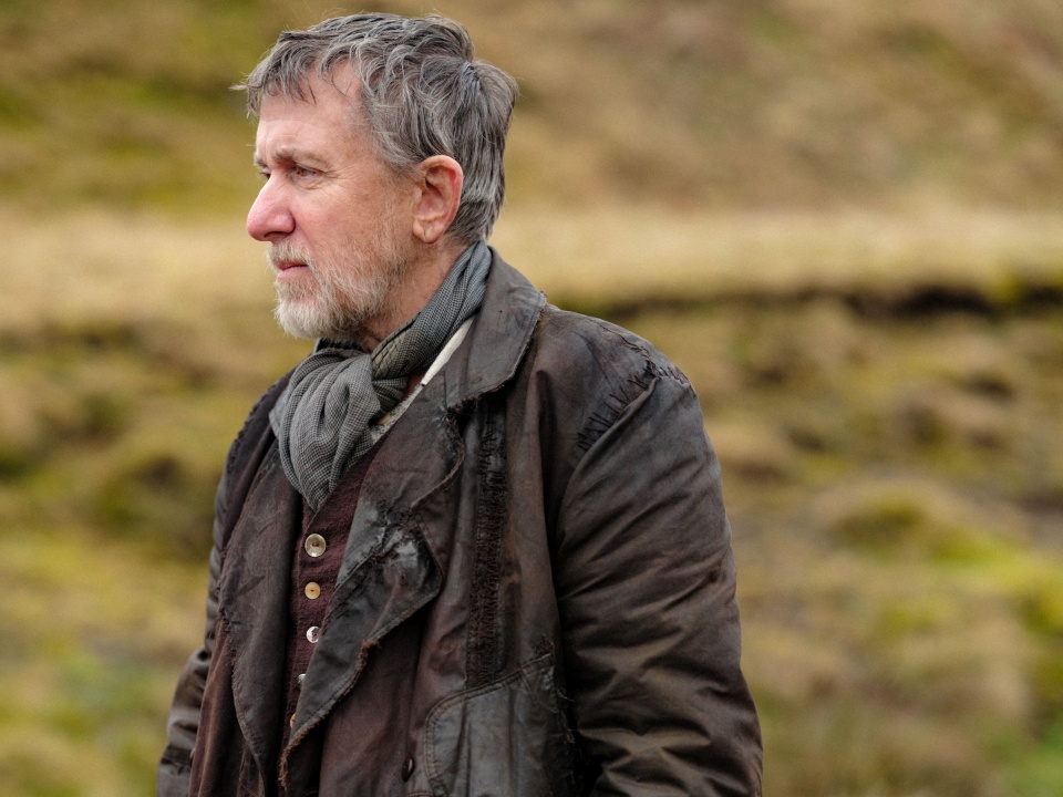 A man with gray hair and a beard, wearing a worn brown coat and scarf, stands outdoors in a grassy, hilly area.