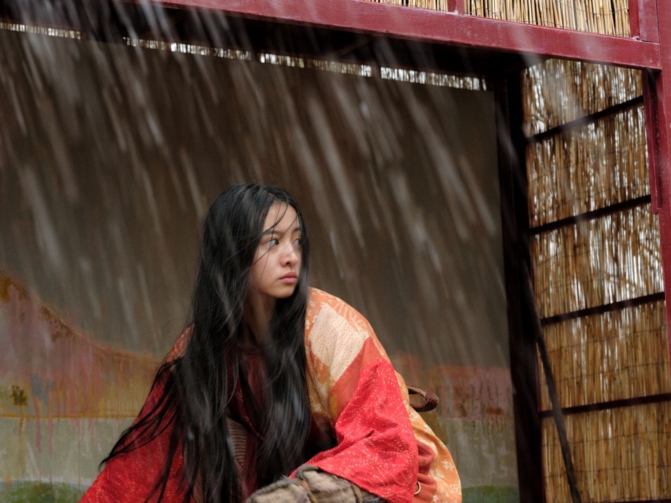 A person with long dark hair wearing a red and orange robe sits under a shelter, looking to the side as rain falls heavily outside.