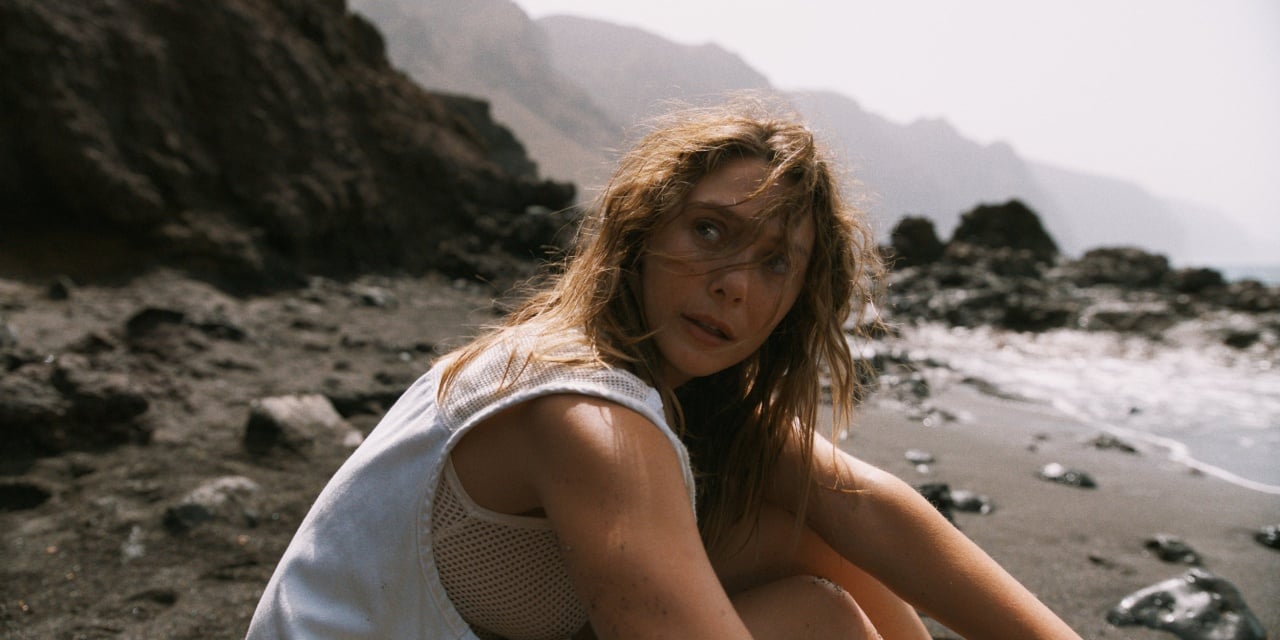 A person with tousled hair sits on a rocky beach near the water, looking towards the camera with mountains in the background.