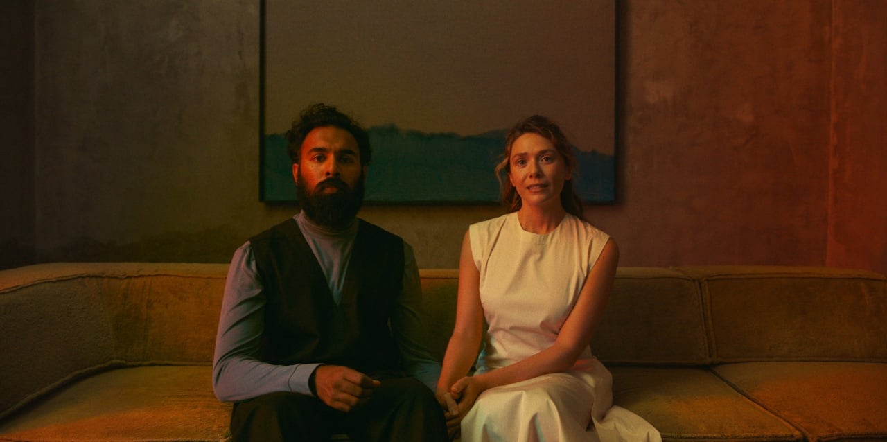 A man in dark clothing and a woman in a white dress sit side by side on a sofa with a painting hanging on the wall behind them.