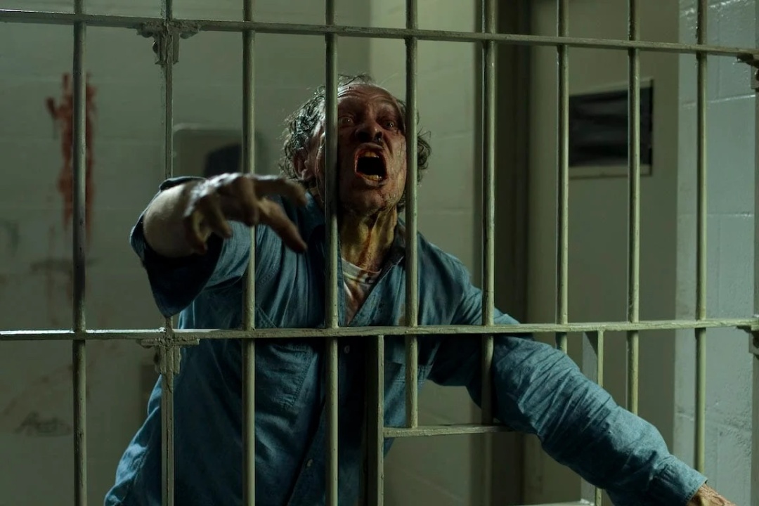 A disheveled man with blood on his face and shirt reaches through the bars of a jail cell, appearing distressed and agitated.