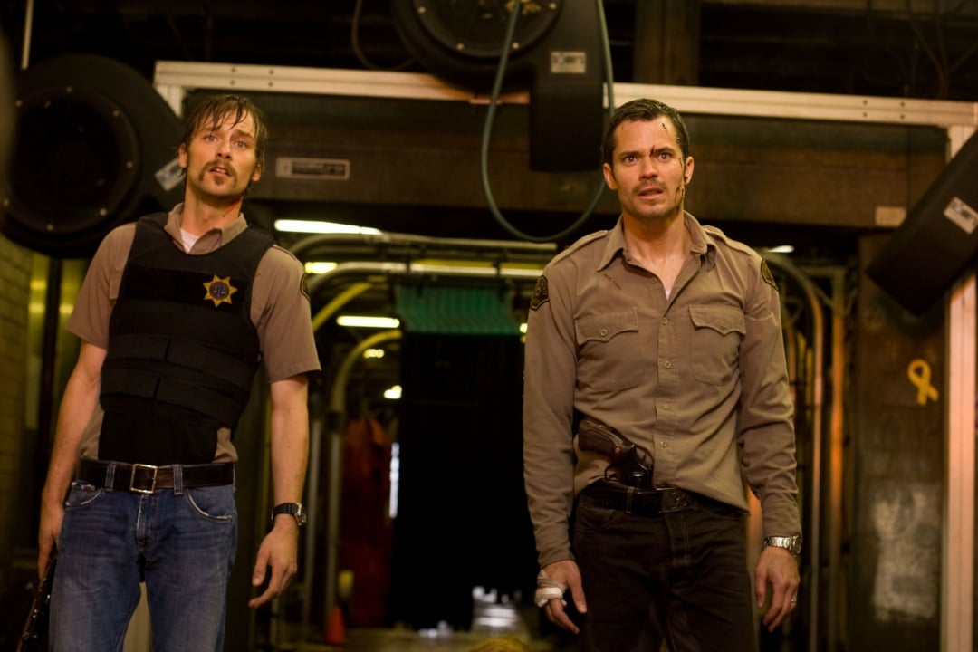 Two men in sheriff uniforms stand alert in a dimly lit industrial setting, both appearing tense and ready for action.