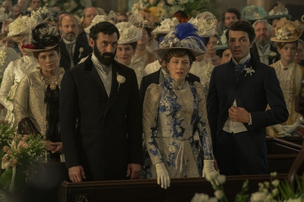 A group of well-dressed men and women in ornate period clothing and hats attend a formal event, standing in a crowded, decorated room.