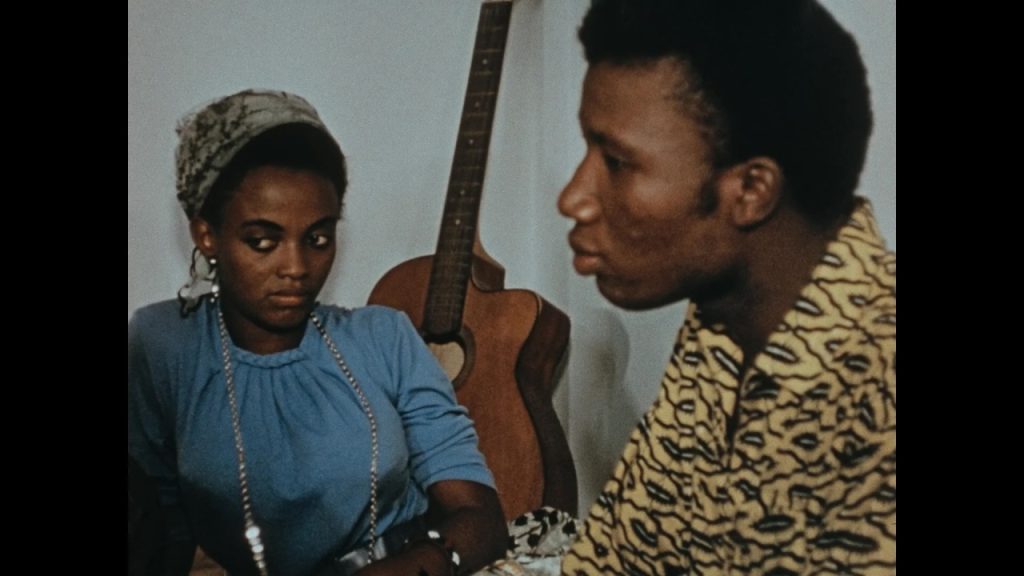 A woman in a blue blouse and headscarf sits next to a man in a patterned shirt; an acoustic guitar rests against the wall behind them.