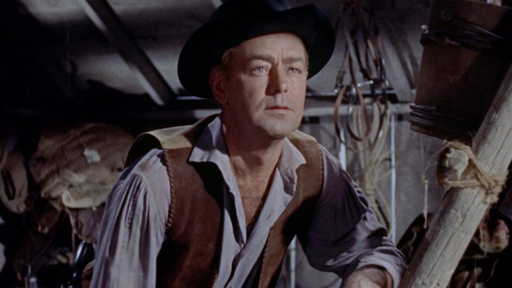 A man wearing a black hat and brown vest sits indoors, looking ahead with a serious expression. Ropes and wooden beams are visible in the background.