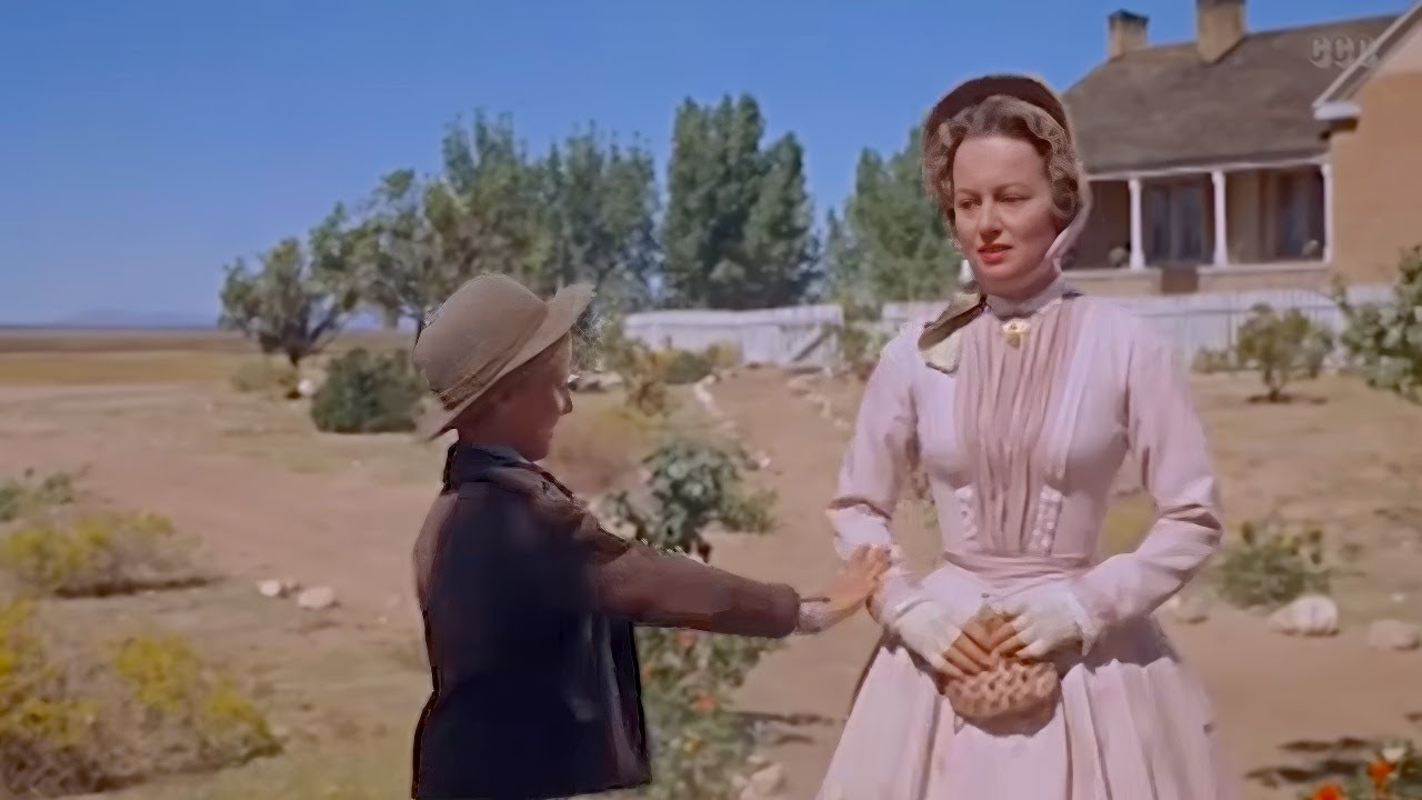A woman in a pale dress and bonnet stands outside near a boy in a brown jacket and hat, with a house and trees in the background under a clear sky.