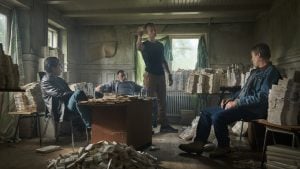 Four men sit and stand in a rundown room filled with large stacks of money piled on tables, desks, and the floor.