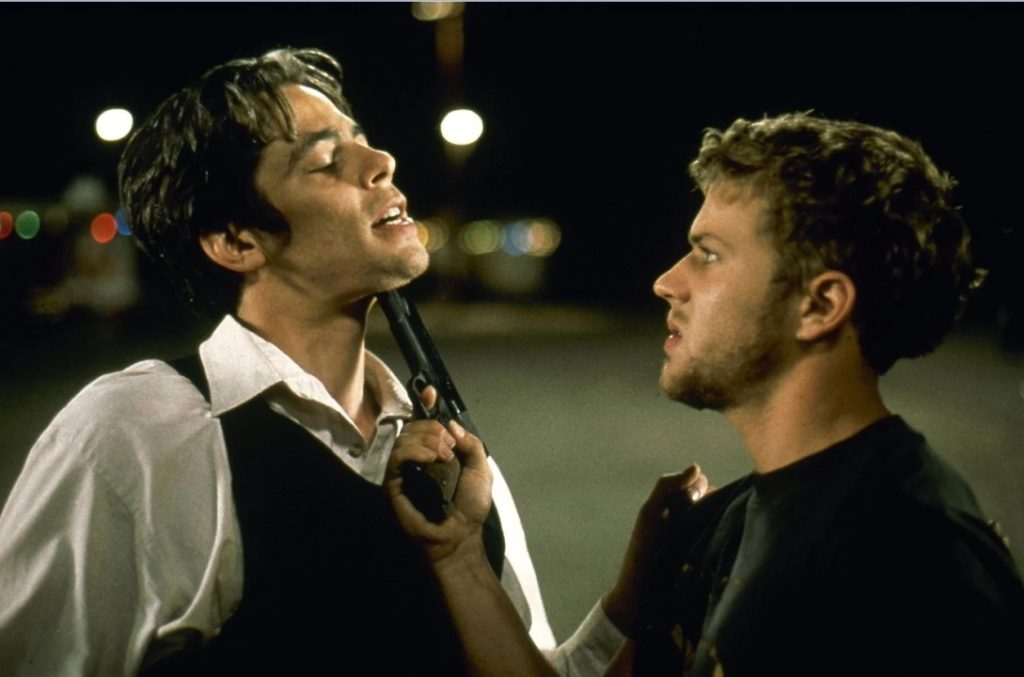 Two men face each other at night; one holds a gun pressed against the other's chin in a tense confrontation.