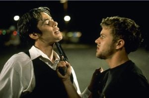 Two men face each other at night; one holds a gun pressed against the other's chin in a tense confrontation.