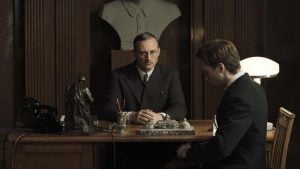 Two men in suits sit across from each other at a wooden desk in an office with dark wood paneling and a bust sculpture on a shelf behind them.