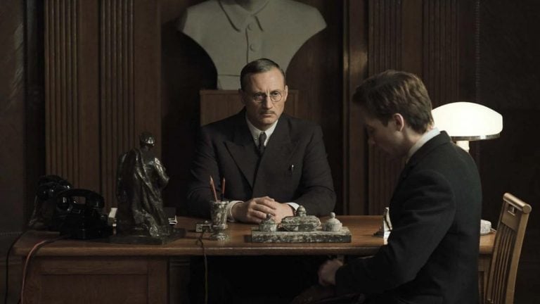 ‘Two Prosecutors’ Criterion Collection Blu-Ray Review – A Captivating Tale Of The Horrors Of Totalitarianism Two men in suits sit across from each other at a wooden desk in an office with dark wood paneling and a bust sculpture on a shelf behind them.