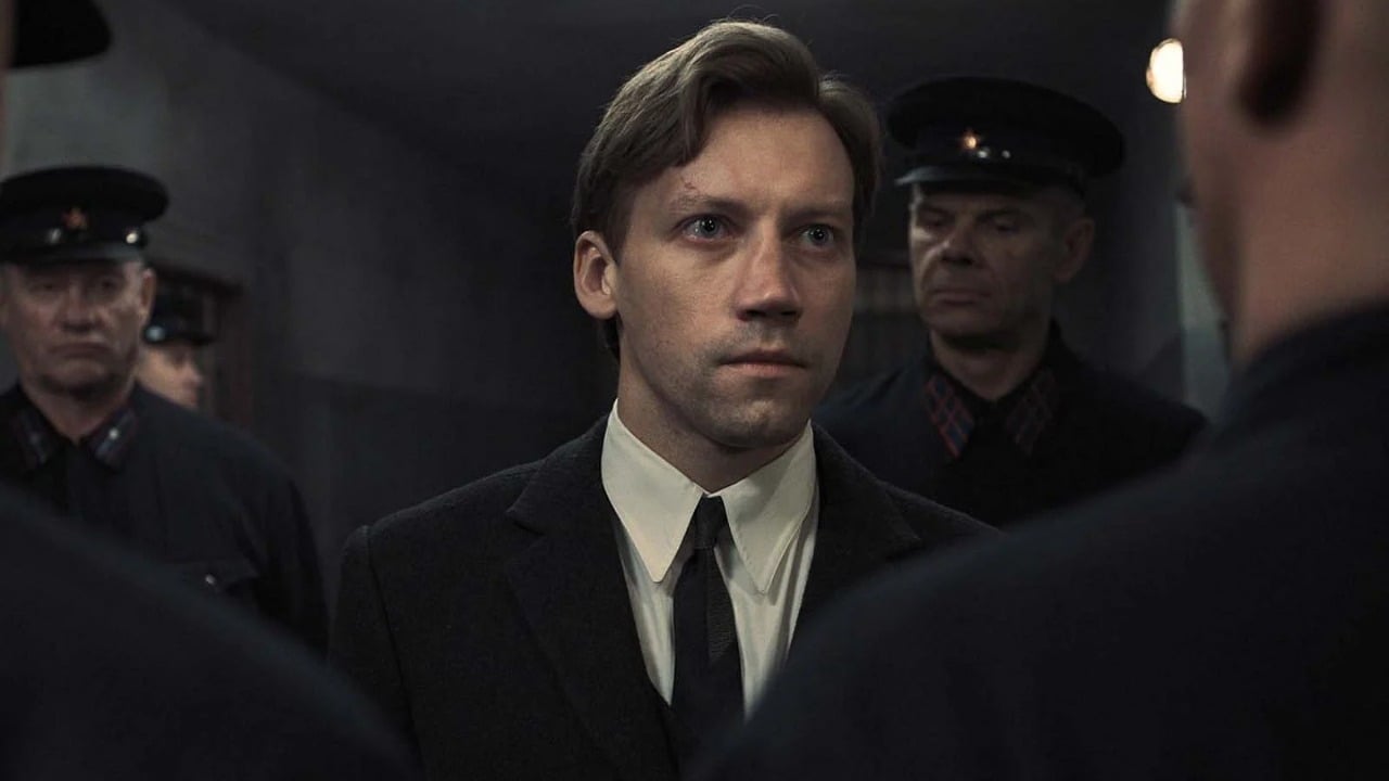 A man in a suit stands in a dim hallway, surrounded by uniformed officers, facing forward with a serious expression.