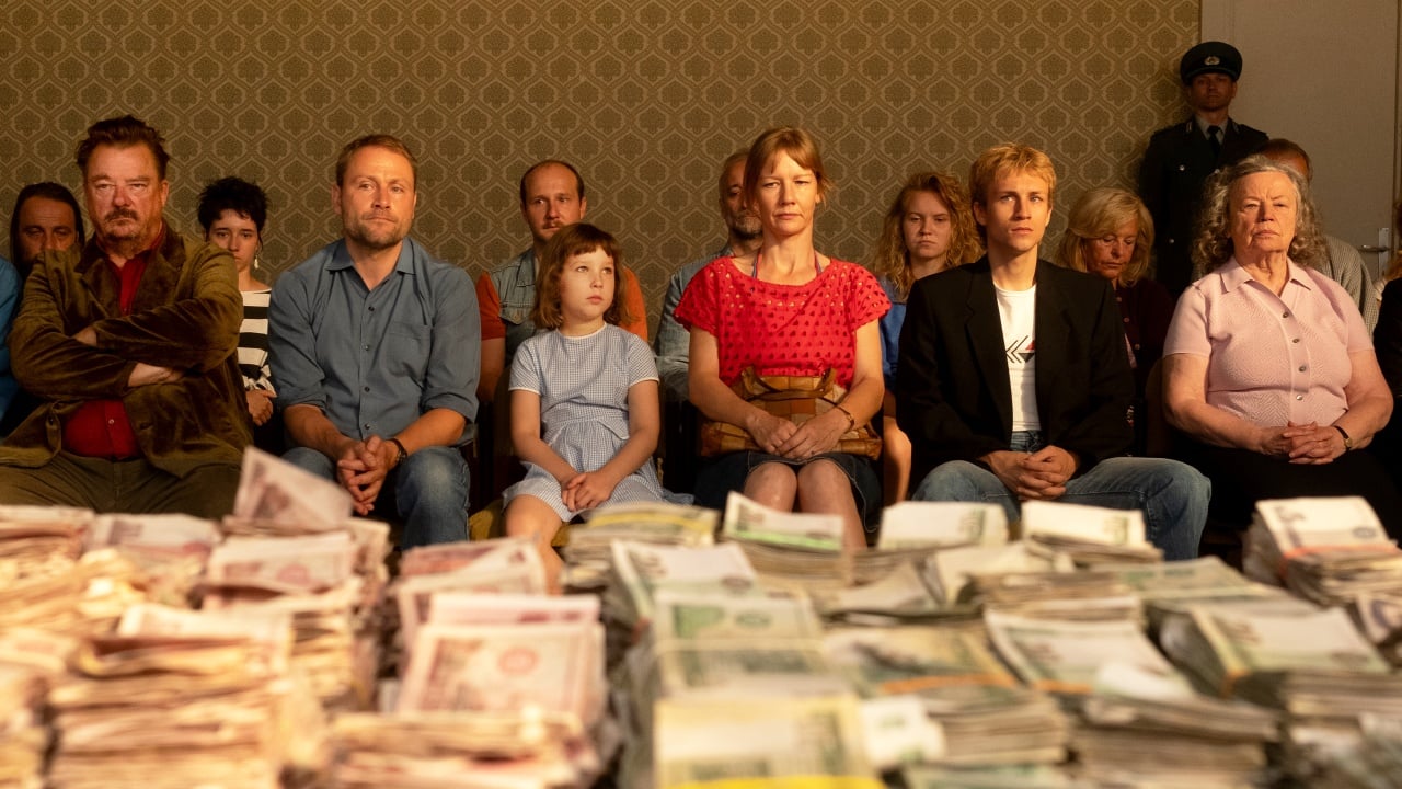 A group of people sit in a row facing stacks of assorted currency on a table, with a patterned wallpaper background and a uniformed guard standing in the back.