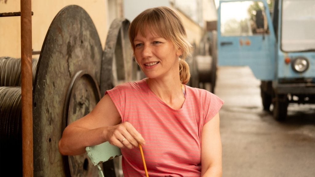 A woman in a pink striped shirt sits outdoors on a chair, holding a pencil, with large spools and a blue vehicle in the background.