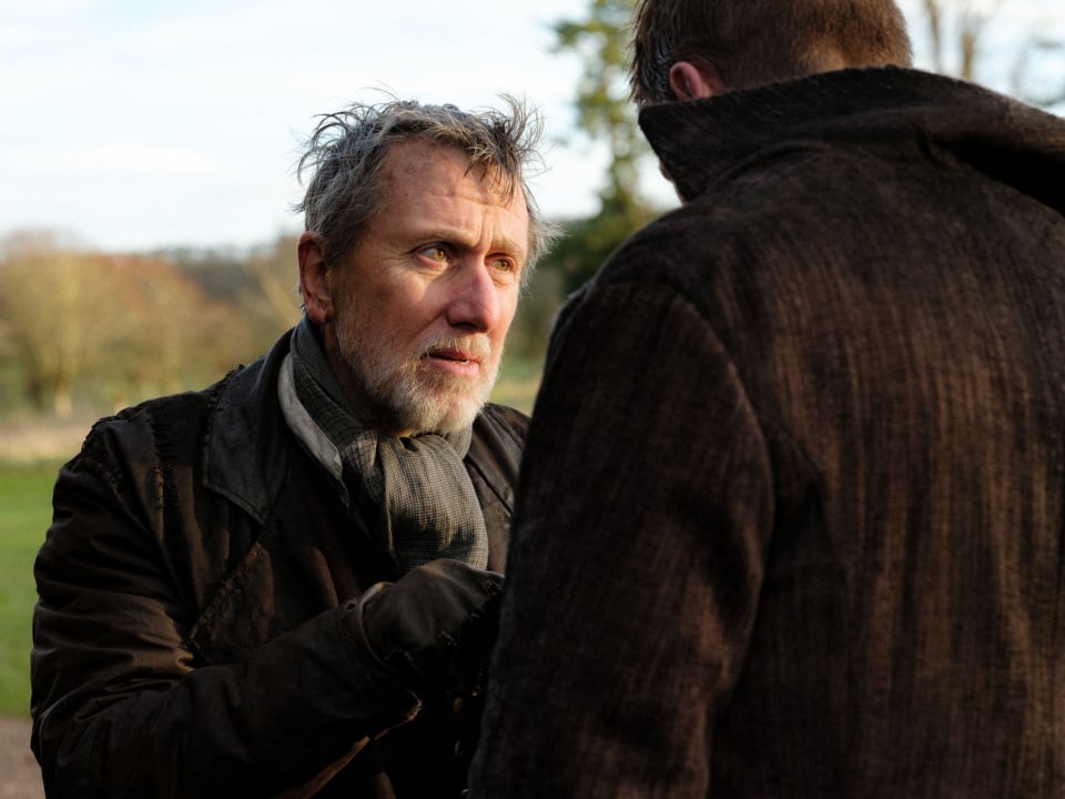 An older man with gray hair and beard, wearing a scarf and coat, stands outdoors talking to another man whose back is facing the camera.