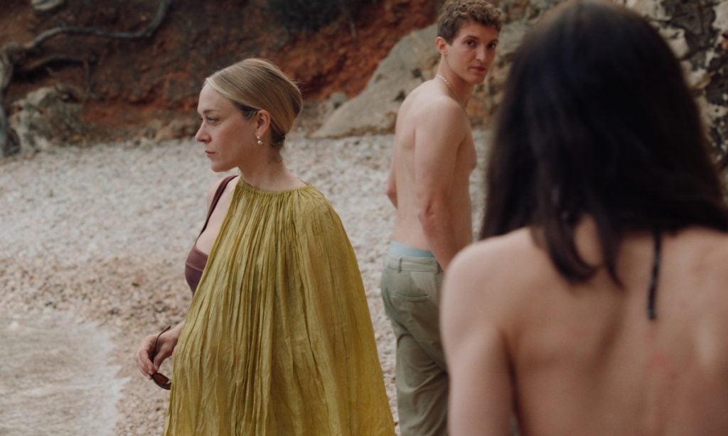 Three people stand outdoors on a rocky path; one woman in a yellow dress faces left, while a shirtless man and a topless person with long hair look toward her.