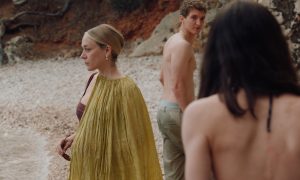 Three people stand outdoors on a rocky path; one woman in a yellow dress faces left, while a shirtless man and a topless person with long hair look toward her.