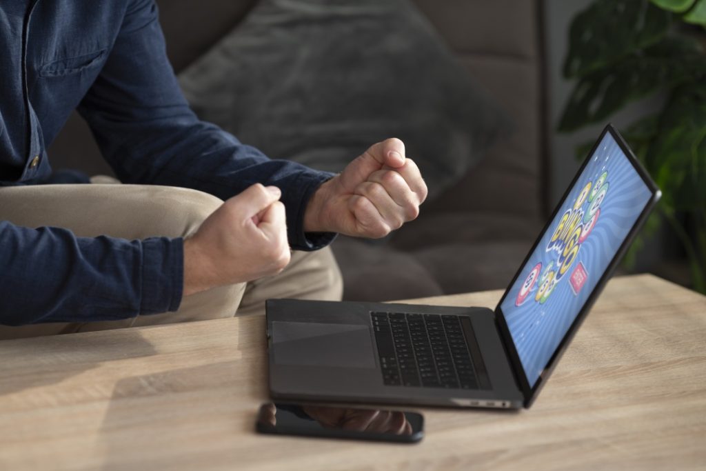 A person sitting at a table clenches their fists in front of a laptop displaying a colorful screen, with a smartphone lying nearby.