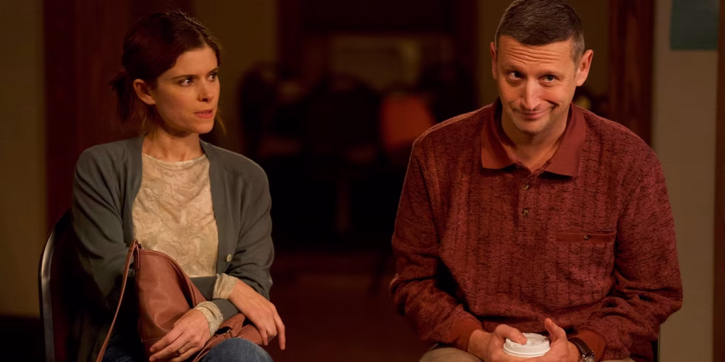 A woman and a man sit side by side indoors; the woman looks skeptical while the man, holding a cup, smiles slightly.
