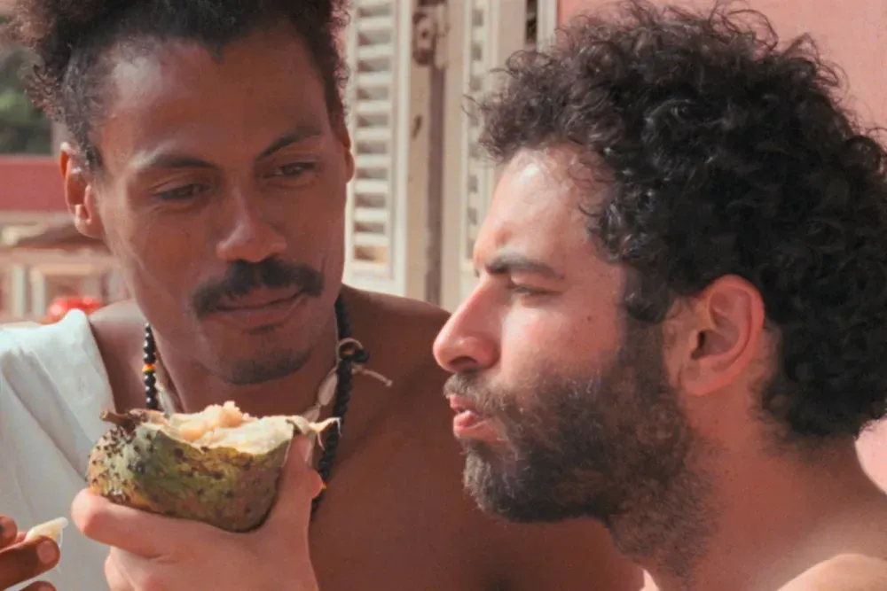 Two men are outdoors; one is holding a piece of fruit while the other, with curly hair and a beard, appears to be eating or tasting it.