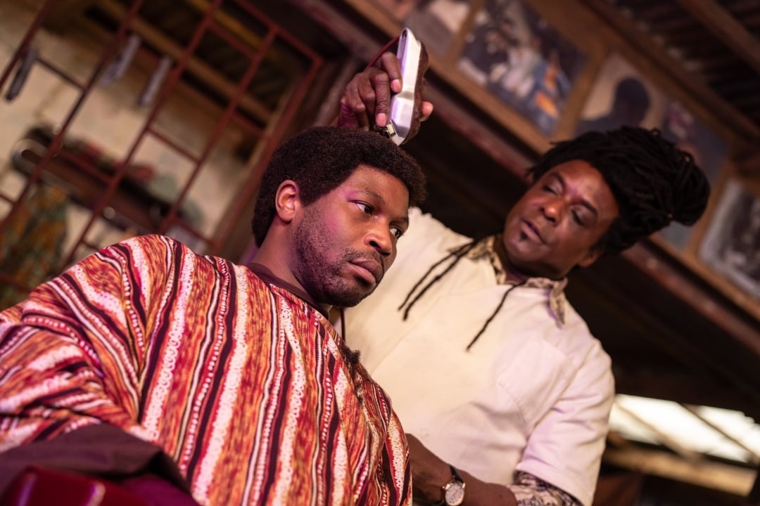 A man sits in a barber’s chair with a striped cape while a barber trims his hair with electric clippers inside a barbershop.
